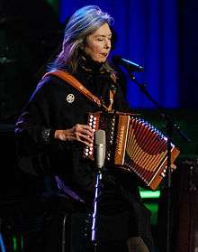 A woman in dark clothing behind a microphone stand on a stage; her eyes are closed, and she is playing an accordion
