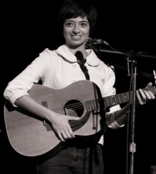 Micucci on stage at microphone, holding guitar, with her trademark bob hairstyle, wearing a bright big-buttoned midi-sleeve blouse