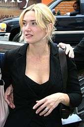 a young woman, with casually styled blonde hair wears a black jacket over a black dress. She is walking along a street; behind her a man sits in a car looking in her direction.