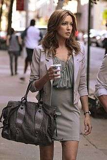 A tall blonde woman carrying a metallic handbag and a coffee cup walks next to an individual wearing a white shirt