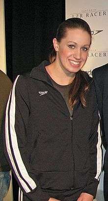 Young woman with long brown hair stands with her hands by her waists and protrudes her head forward. She is wearing a black Speedo tracksuit which is partially zipped up, and an olive shirt underneath.