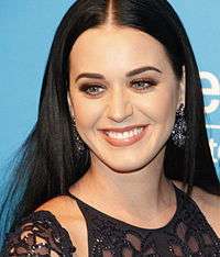 A woman with pale skin, green eyes and a long dark hair and wearing a dark dress smiles to the camera.