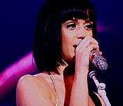Katy Perry with short, black hair singing into a silver sparkly microphone.