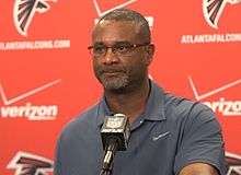 Candid chest-up photograph of Armstrong standing at a podium with Atlanta Falcons logos on the backdrop during a press conference wearing a teal polo shirt with a Nike logo and eyeglasses.