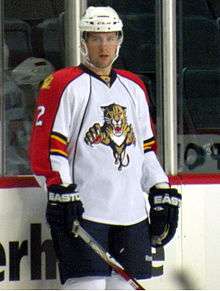A Caucasian ice hockey player standing relaxed on the ice. He wears a white, visored helmet and a white and orange jersey. He holds his stick on the ice lightly with one hand.