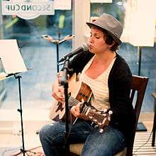 A photograph of a woman wearing a grey hat, a yellow-and-white striped shirt, and blue jeans playing a guitar and singing into a microphone