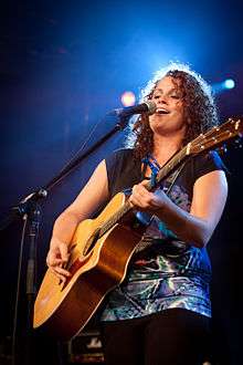 A photograph of a woman with shoulder-length, curly, brown hair playing a guitar with a pick and singing into a microphone, backlit with a blue light