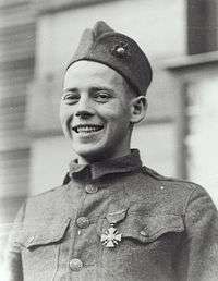 Head and shoulders of a happy young man in a military uniform with a single medal on his chest. He is smiling and his garrison cap is pushed high up on his forehead.
