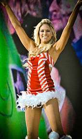 A blonde, Caucasian woman smiles while raising both her hands above her head. She is wearing a red and white dress with a white fur trim.