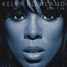 A close-up image of a woman's face covers the image. Her skin is blue and she is looking directly at the camera, with her face framed by her long black hair. Across the top in white text it reads KELLY ROWLAND (the name of the artist). On the line immediately below, in smaller text and aligned to the right, it reads HERE I AM (the name of the album).
