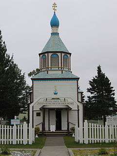 Photograph of the front of the Holy Assumption Orthodox Church, behind a white picket fence with white clapboard and a blue roof and onion dome.