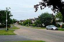A photograph of a quiet suburban street. The houses are large and semi-detached, fronted by paved driveways and mown grass verges.