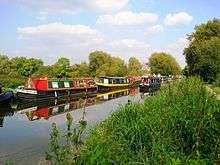 A canal passing through countryside. Numerous narrowboats are moored on the far bank, and a narrowboat is travelling upstream towards the foreground.