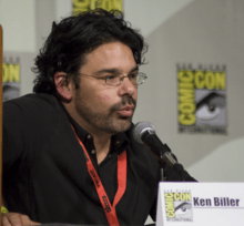 A bearded, dark-haired man leans into a microphone; he is sitting behind a San Diego Comic Con paper placard that declares his name "Kenneth Biller".