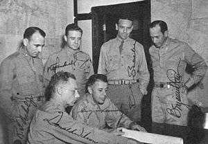 Kenney and Wilson sit at a table. Four other men in uniforms look on with their hands in their pockets. Each has signed the photograph.