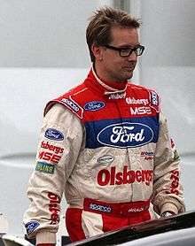 Man in his mid forties, wearing white, blue and red racing overalls. He has a head full of hair and is wearing glasses.