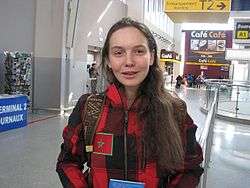 A woman with long brown hair faces the camera, she wears a red and black checked jacket.