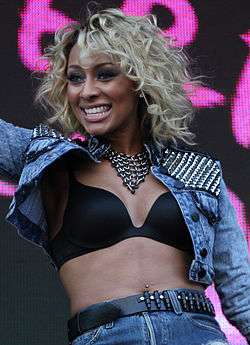 A blonde woman with dark skin, wearing a blue jean jacket and blue short jeans.