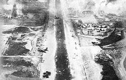 Black and white image from the rear of a plane showing a landing strip littered with damaged aircraft