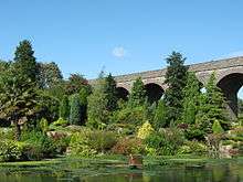 View of a garden containing a number of evergreen trees and a palm tree, with a lake in the foreground and the arches of a disused railway viaduct in the background.