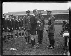 A black and white photo of a rugby field in which three men in military uniform, one of whom is King George, present a silver trophy to a rugby player dressed in black kit. Behind in a line are the rest of the team.