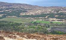 A view from an elevation above cultivated fields and a mixed wood of conifers and deciduous trees. Various indistinct buildings are visible among these trees. The brown ridge of a hill in the background has conifers growing on its lower slopes.