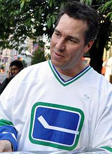 A middle-aged, Caucasian man wearing a white ice hockey jersey with a blue and green logo of a horizontal hockey stick.  His head is tilted and he is looking towards the right.  His hair is dark brown and spiked upwards in the front.