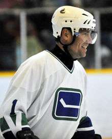 A side view of a middle-aged, Caucasian ice hockey player. He is wearing a white helmet and jersey with a blue and green logo of a horizontal hockey stick. He looking forward and smiling.