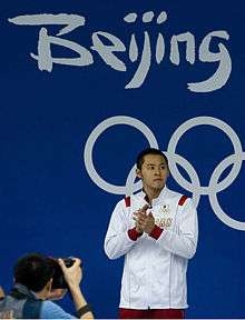 Asian man with short black hair, in white tracksuit stands on the podium waiting to receive his medal. Behind him is a blue wall with the words "Beijing" and "2008" in white.