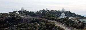 Panoramic view of a mountain top with trees and some white domed telescope buildings and a road leading up to the top.