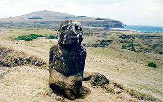 colour photo of kneeling figure carved from red stone, head splashed with white. A triangular hill is in the background and the sea in the top right