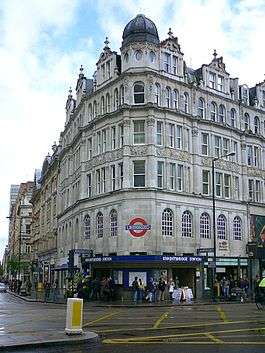 A white building with a rectangular, dark blue sign reading "KNIGHTSBRIDGE STATION" in white letters all under a light blue sky with white clouds