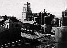 A black-and-white photo of the shale oil processing facility at Kohtla-Järve, dated to 1937. A rail line is shown in the lower third of the photo. A limestone-walled rock hopper tower building and a generator-house are located in the background. Another smaller building is located in the left side of the photo. Several oil tanks are located in the right side of the photo.