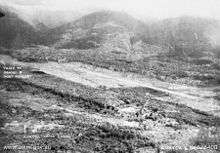 Panoramic view of a cleared strip of land surrounded by thick scrub and jungle. Behind the cleared strip the ground rises steeply