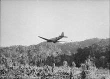 A transport plane flying at low level away from the camera, dropping supplies over a clearing in the jungle