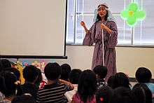 A photograph of a woman wearing a red-and-blue-striped robe holding a white stick and speaking while standing in front of a group of children who are facing her