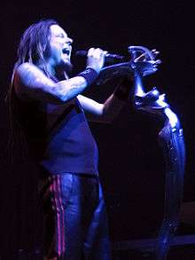 A man with his eyes closed and mouth open, holding a microphone; he is wearing dark clothing and wrist bands.