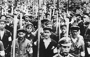 A black and white picture of a large group of men marching towards the camera, most of them dressed in civilian clothes and armbands, presumably in colours of the Polish national flag. The men carry long war scythes. To their right a large crowd of civilians.