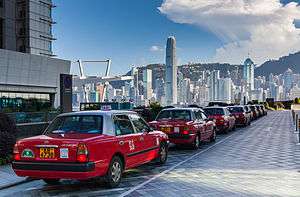 Taxis lined up along a drive outside a building. In the background is a city skyline with tall skyscrapers and a ridgeline under a blue sky with some clouds.