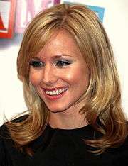 Head and shoulders of young, blond woman with hair curling below her shoulders, smiling.