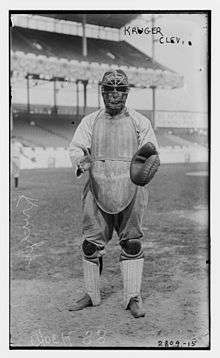 A man wearing a baseball uniform and full catcher's gear (facemask, catcher's mitt, chest protector, and leg guards) stands poised to catch a ball.
