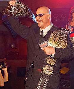 Adult white male wearing a black suit and sunglasses holding two black belted championship belts and a microphone.