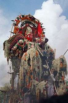 A huge sculpture of an armoured warrior man, with a moustache and red face. He is adorned with many floral garlands, which also cover his arms. Male priests surround him.