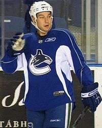 Hockey player in blue uniform, with a "C" in the middle. One of his hands is raised, and the other grasps his hockey stick.