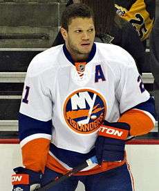 Kyle Okposo standing on the ice with his hockey stick and no helmet on.