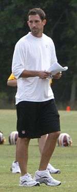 Candid photograph of Shanahan wearing a white long-sleeved t-shirt and black shorts and standing on a football practice field holding a small sheaf of papers in his hands