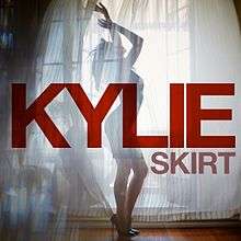 Minogue standing in heels behind a curtain, with the words 'Kylie' and 'Skirt' superimposed in a red font.