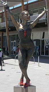 A bronze statue of Kylie, on a star-shaped pedestal, portrays her in a dancing pose. Her legs are crossed and she bends at the waist, with both arms stretched above her head. The statue stands in a public square in front of a modern glass building, and several people are walking.
