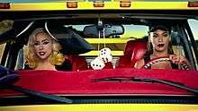 Gaga sitting beside Beyoncé who is driving a yellow colored van. Gaga wears a giant hat on her head. A pair of dice hangs from the rear-view mirror between them.