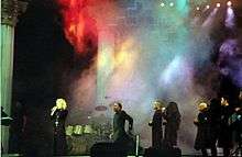 Faraway image of a female blond singer wearing a black robe with a crucifix and singing to a microphone she holds in her hand. Behind here several African-American dancers dressed similarly can be seen. The staged is filled with a colored smoke and some drums and a cross can be seen.